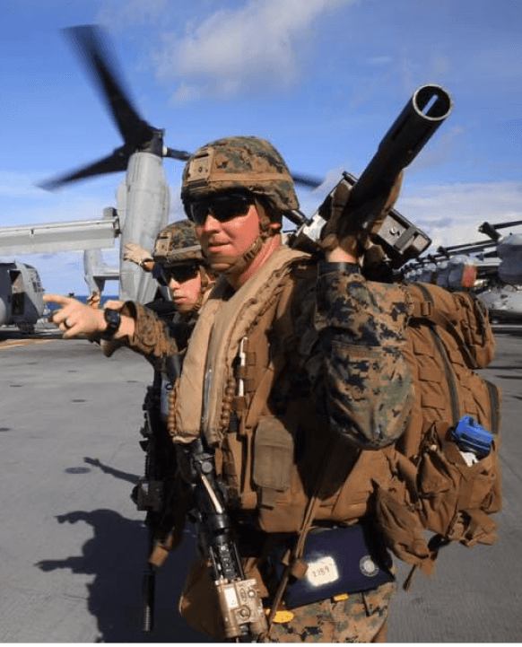 Zach on his second deployment. He's standing on a Ridgeline in the Australian Outback in his camouflage uniform wearing body armor and war paint, and has an M240B medium machinegun slung over one shoulder. He's standing next to another machine gunner and with the coastline in the distant background.