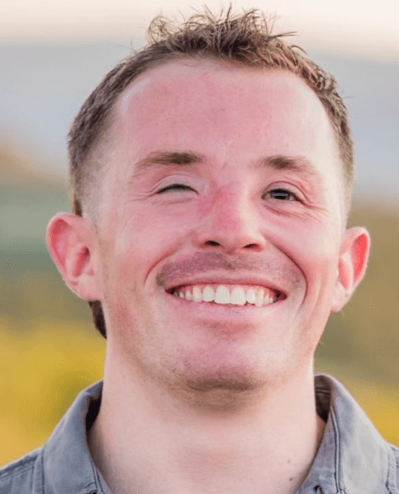 A close-up of Zach since losing his sight. He has light brown hair, blue eyes, and a big smile. He has some scarring between his eyes and across the bridge of his nose from his facial reconstruction surgery, and his right eye does not open all the way.